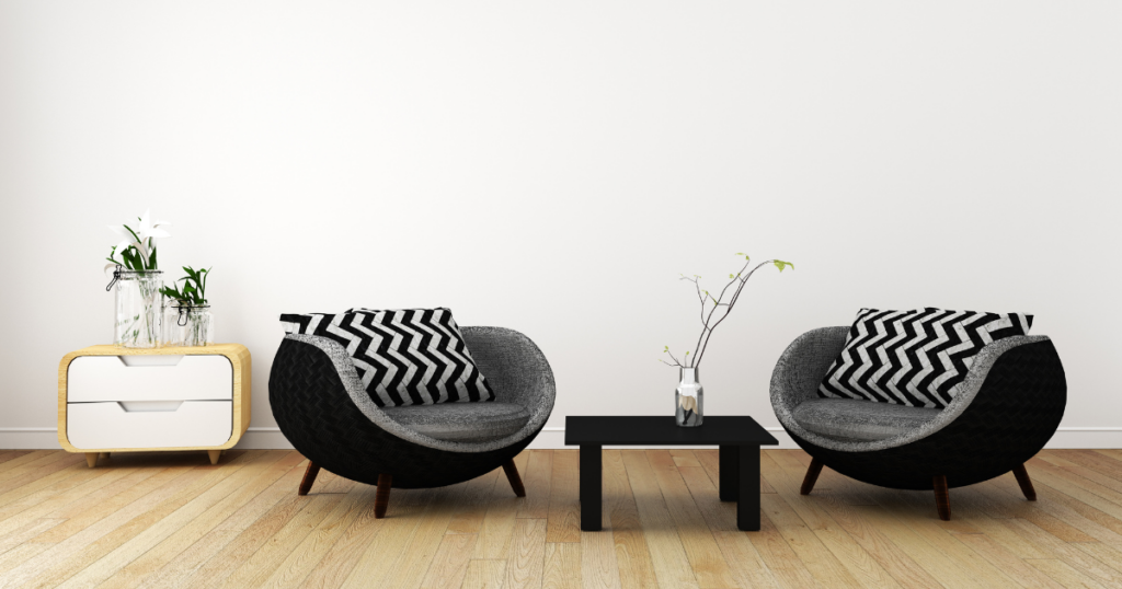 This room, thoughtfully modified for vision loss, is not only aesthetically pleasing but also highly functional. The white walls and light wood floors provide a bright, open space, while the two-drawer dresser, adorned with two green plants, offers a practical storage solution. To the right, two black chairs with black and grey zigzag pillows provide comfortable seating, and a black coffee table with a plant sitting in a vase serves as a convenient surface for everyday use. 