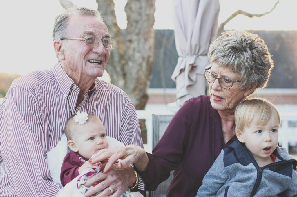 A caucasian heterosexual elderly couple is holding their grandchildren. The grandfather on the left is wearing a pink and white vertical-striped shirt. On his lap is a baby wearing a white flower on her head and a maroon dress. To the left is a grandmother, wearing dark-framed glasses and a maroon v-neck shirt. A young boy with blonde hair is on her lap, wearing a black and grey jacket.