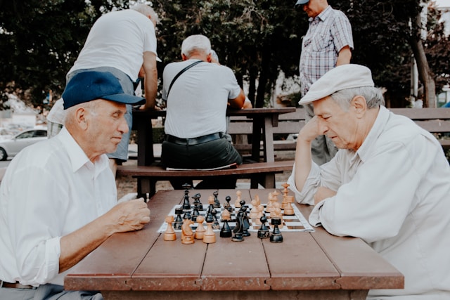 Two older Caucasian men are playing chess outside in a senior living community. They are sitting facing each other at a wooden table. The man on the left wears a blue baseball hat and a white button-down shirt. The man on the right is wearing a white cloth baseball hat and a white button-down shirt.