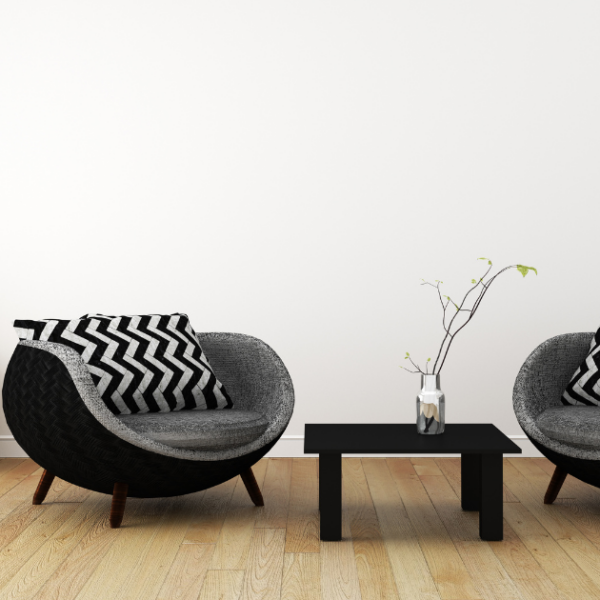 This room, thoughtfully modified for vision loss, is not only aesthetically pleasing but also highly functional. The white walls and light wood floors provide a bright, open space, while the two-drawer dresser, adorned with two green plants, offers a practical storage solution. To the right, two black chairs with black and grey zigzag pillows provide comfortable seating, and a black coffee table with a plant sitting in a vase serves as a convenient surface for everyday use.