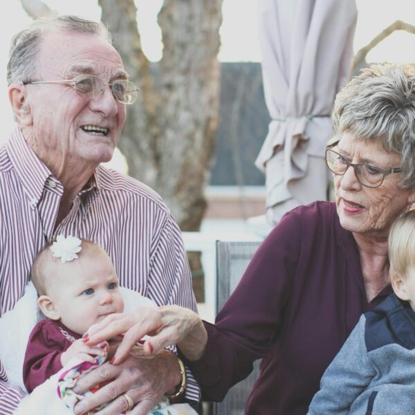 A caucasian heterosexual elderly couple is holding their grandchildren. The grandfather on the left is wearing a pink and white vertical-striped shirt. On his lap is a baby wearing a white flower on her head and a maroon dress. To the left is a grandmother, wearing dark-framed glasses and a maroon v-neck shirt. A young boy with blonde hair is on her lap, wearing a black and grey jacket.