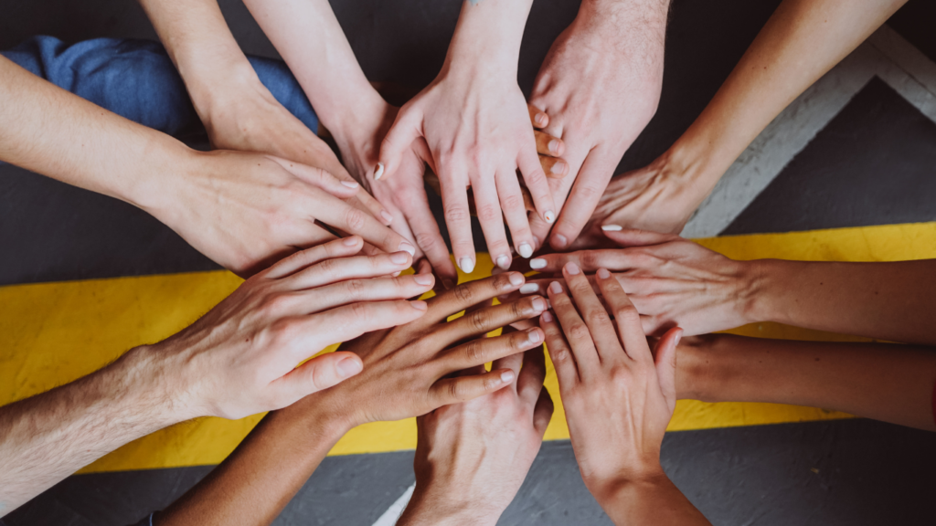 Twelve hands of various skin tones are on each other to show support.