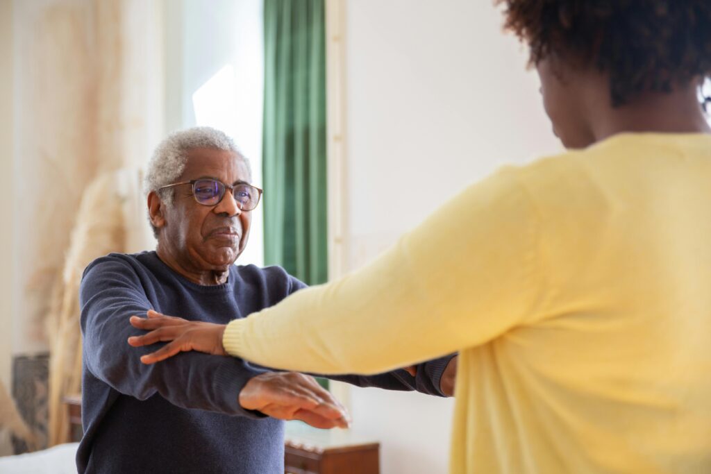 An older male in glasses and a long-sleeved blue shirt face a younger woman in an afro and yellow cardigan. She is helping him do physical exercise, possibly in memory care.