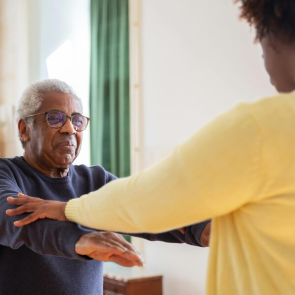 An older male in glasses and a long-sleeved blue shirt face a younger woman in an afro and yellow cardigan. She is helping him do physical exercise, possibly in memory care.